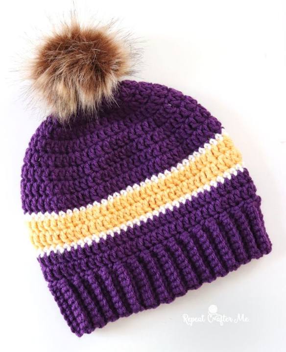Purple and gold striped crochet beanie with a ribbed brim and fur pom pom