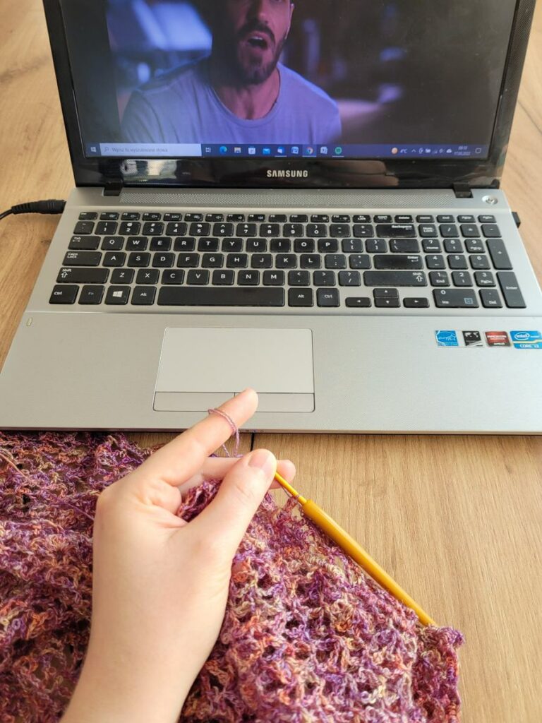 Crocheting a lace shawl in pink and purple yarn in front of a laptop